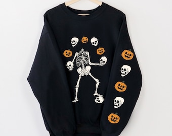 May include: Black Halloween sweatshirt featuring a skeleton juggling skulls and pumpkins. The design includes orange pumpkins and white skulls on the sleeves and around the skeleton. The word "Black" is printed at the bottom left.