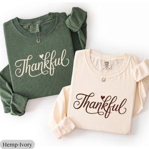 May include: Two long-sleeved shirts, one in olive green and the other in ivory, are displayed. The word "Thankful" is written in a cursive font on the front of each shirt. Each shirt has a small pendant necklace.