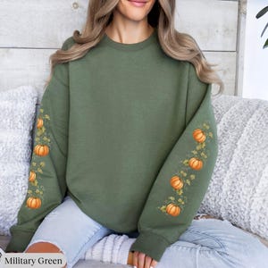 May include: A military green crewneck sweatshirt featuring long sleeves decorated with a repeating graphic print of orange pumpkins and green vines running down the length of each sleeve. The garment has a ribbed collar, cuffs, and hemline.
