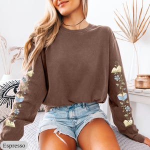 May include: A brown long-sleeve crewneck sweatshirt featuring a graphic print on both sleeves. The design consists of winding green vines with small blue flowers and open books interspersed along the length of each sleeve.