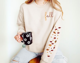 May include: A sand-colored sweatshirt with the word "fall" in script on the chest. The right sleeve features a design of falling leaves in shades of brown and orange. A black mug with white flower designs is held in the left hand.