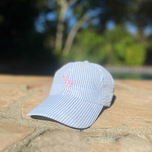 Könnte beinhalten: Eine blaue und weiße gestreifte Baseballkappe mit einem rosa gestickten Monogramm.