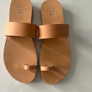 May include: A pair of tan leather sandals with a toe loop and a single strap across the top of the foot. The sandals have a leather sole and are marked "MADE IN GREECE" on the bottom.
