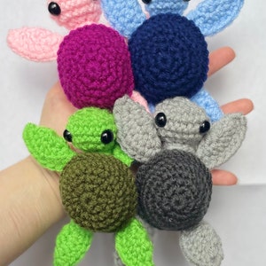 May include: Five crocheted sea turtle toys in different colors: pink, blue, purple, gray, and green. Each turtle has black eyes and a crocheted shell.