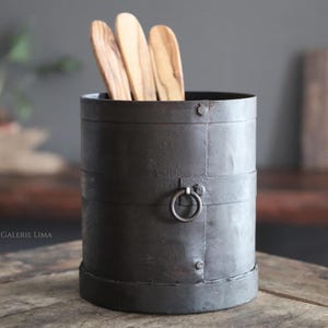 Könnte beinhalten: Ein schwarzer Metall-Besteckhalter mit einem Ringgriff und Holzlöffeln im Inneren. Der Halter hat ein rustikales, gealtertes Aussehen.