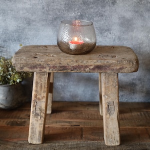 Könnte beinhalten: Ein rustikaler Holzhocker mit einer verwitterten Oberfläche, auf dem ein kleiner Glas-Kerzenhalter mit einer brennenden Kerze steht. Der Hocker steht vor einer strukturierten grauen Wand.