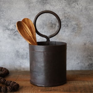 Op de afbeelding: Een donkerbruine metalen keukengerei houder met een ronde handgreep, met twee houten lepels. De houder is cilindrisch en staat op een houten oppervlak. Een streng bruine kralen staat op de voorgrond.