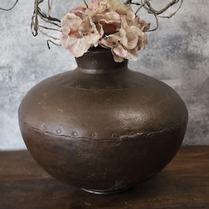Puede incluir: Un jarrón redondo de metal marrón oscuro con una boca ancha, decorado con un patrón de pequeños círculos. El jarrón está lleno de flores rosadas secas.