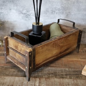 Könnte beinhalten: Eine rustikale Holzkiste mit Metallverzierungen und Beinen. Die Kiste ist gefüllt mit einer schwarzen Glasdiffusor-Flasche mit goldenen Akzenten und einem grünen Dekorationsgegenstand.