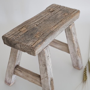 Könnte beinhalten: Ein kleiner, rustikaler Holzschemel mit rechteckiger Sitzfläche und schrägen Beinen. Das Holz hat ein verwittertes, graubraunes Aussehen mit sichtbarer Maserung und Knoten. Der Hocker ist etwa 30 cm lang, 20 cm breit und 25 cm hoch.