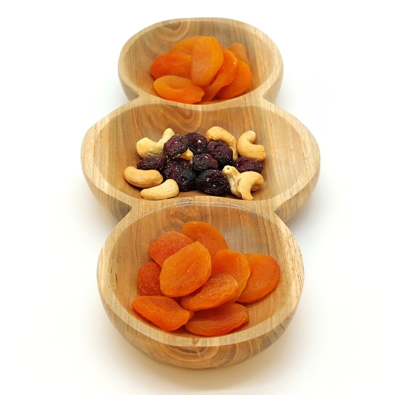 May include: A wooden serving dish with three separate bowls. The top bowl contains dried apricots, the middle bowl contains dried cranberries and cashews, and the bottom bowl contains more dried apricots.