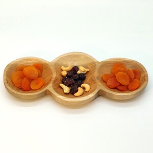 May include: A wooden serving dish with three separate compartments. The compartments are filled with dried apricots, cashews, and cranberries.
