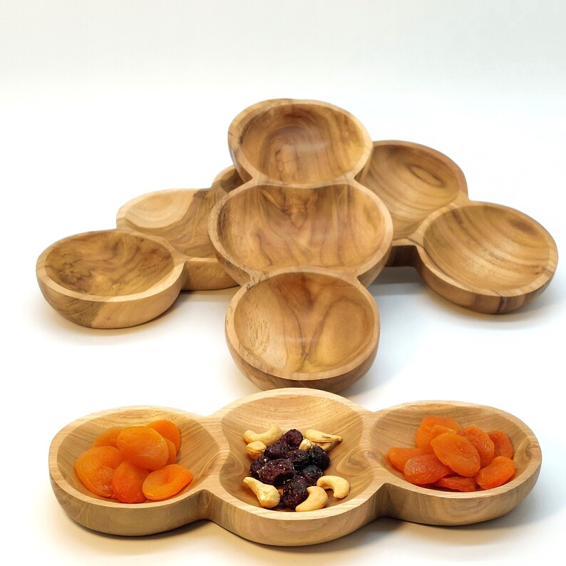 May include: A wooden serving tray with three separate bowls. The bowls are filled with dried apricots, cashews, and cranberries.