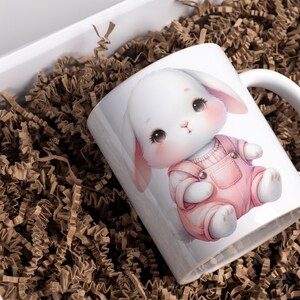 K&ouml;nnte beinhalten: Wei&szlig;e Keramiktasse mit einer Cartoon-Illustration eines wei&szlig;en Kaninchens, das eine rosafarbene Latzhose tr&auml;gt. Das Kaninchen hat gro&szlig;e, schwarze Augen und eine rosafarbene Nase.