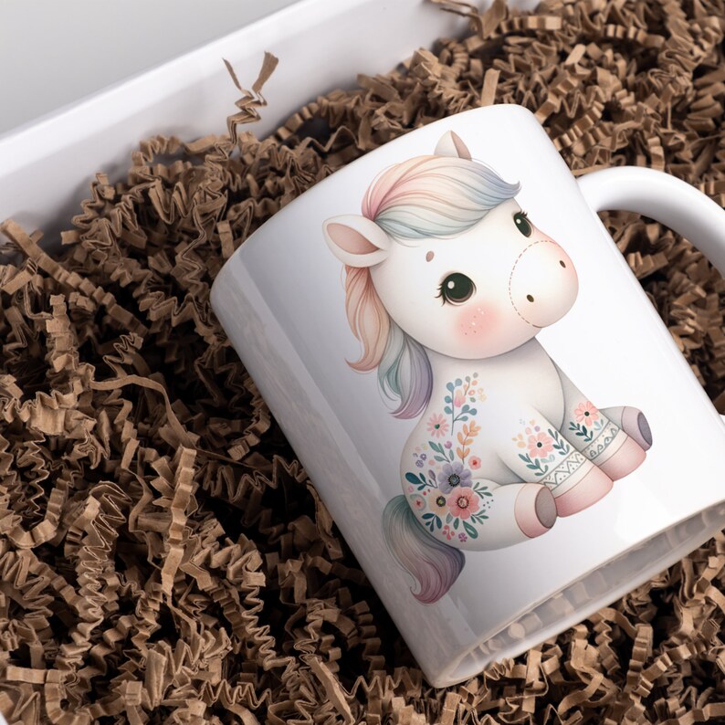 May include: White ceramic mug with a colorful unicorn illustration. The unicorn has a rainbow mane and tail and is adorned with floral designs. The mug is sitting in a brown paper shred.