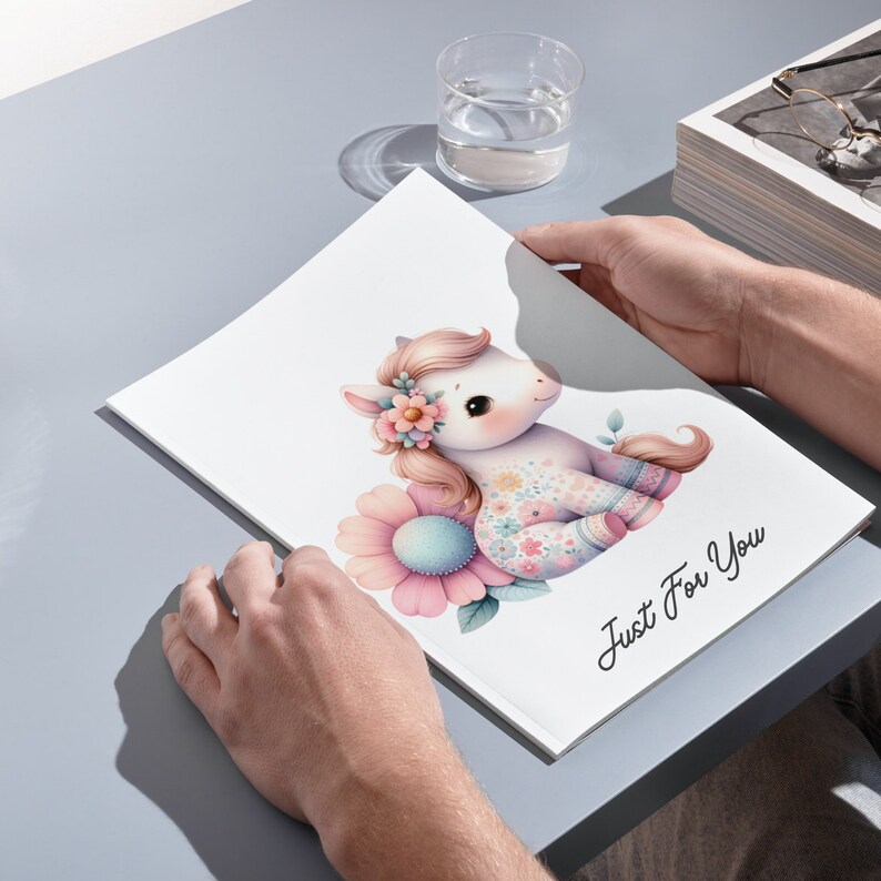 May include: A white notebook with a pink and blue floral design featuring a cartoon unicorn with a flower crown. The notebook has the text "Just For You" printed on the front.