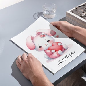 K&ouml;nnte beinhalten: Eine Aquarell-Illustration eines rosa Kaninchens, das ein rotes Herz h&auml;lt. Das Kaninchen tr&auml;gt einen rosa Pullover und hat eine Blume auf dem Kopf. Der Text "Just For You" steht unter dem Kaninchen.