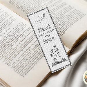 Può includere: Un segnalibro bianco e nero con un design semplice che presenta un libro, un fiore e un aeroplano di carta. Il segnalibro ha il testo "Read between the lines" stampato su di esso.