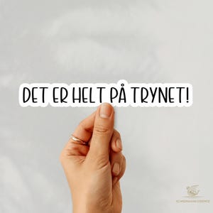 May include: A white sticker with black text that reads "DET ER HELT PÅ TRYNET!". The sticker is held by a hand with a gold ring. The background is a light grey colour.