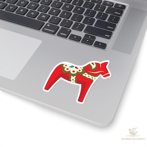 K&ouml;nnte beinhalten: Ein roter Dala-Pferd-Aufkleber mit wei&szlig;en R&auml;ndern, verziert mit gr&uuml;nen, wei&szlig;en und blauen Blumenmustern. Der Aufkleber befindet sich auf einem silbernen Laptop. Das Logo "Scandinavian Essence" ist unten rechts sichtbar.