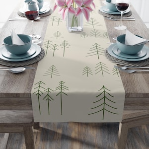 May include: A beige table runner with a repeating pattern of green stylized pine trees. The runner is placed on a wooden table set for a meal, with plates, bowls, silverware, and wine glasses.