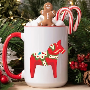 Może przedstawiać: Biały ceramiczny kubek z czerwonym uchwytem i wnętrzem, ozdobiony czerwonym wzorem konia Dala. Kubek wypełniony jest piankami marshmallow, piernikowym ludzikiem i dwoma laseczkami cukrowymi. Świąteczny motyw.