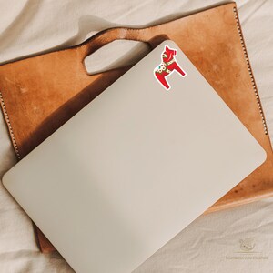 K&ouml;nnte beinhalten: Ein roter und wei&szlig;er Dala-Pferde-Aufkleber mit gr&uuml;nen und goldenen Akzenten befindet sich auf einem geschlossenen, silbernen Laptop. Der Laptop liegt auf einer braunen Ledermappe. Der Hintergrund ist ein cremefarbener Stoff.