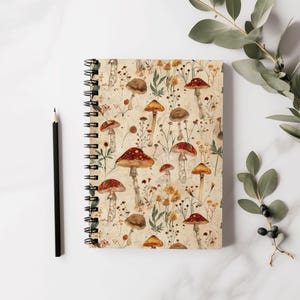 Può includere: Un quaderno a spirale con una copertina color crema che presenta un motivo ripetitivo di funghi rossi e bianchi e fogliame verde.
