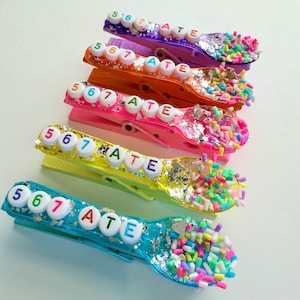 May include: A collection of colorful, decorated clothespins. Each pin is a different color, including purple, orange, pink, yellow, and teal. The pins are adorned with the numbers "5 6 7" and the word "ATE" in white letters, along with glitter and colorful sprinkles.