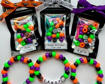 May include: Three black pouches containing colourful Halloween-themed pony beads, each topped with a decorative ribbon. Below are three matching elastic bracelets featuring orange, purple, green, and black beads with letter beads spelling BOO, CANDY, and EEK.