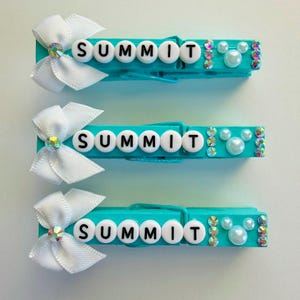 May include: Three turquoise clothespins with the word "SUMMIT" spelled out in white letter beads. Each pin has a white bow with a rhinestone and is decorated with pearls and rhinestones.