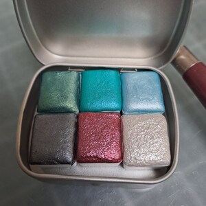 Peut inclure: Une boîte en métal contenant six godets de peinture aquarelle. Les godets sont disposés en deux rangées de trois. Les couleurs sont turquoise, vert, bleu, gris, rouge et argent.