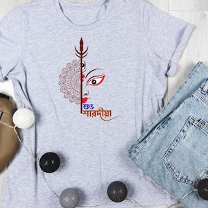 May include: A light grey t-shirt with a graphic of a deity's face, featuring a red eye and brown hair, and a decorative pattern. The Bengali text "শুভ শারদীয়া" is displayed. The shirt is shown with blue denim jeans.