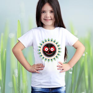 May include: A white t-shirt featuring a colorful graphic design. The design includes a central face with a smiling expression, surrounded by a circular pattern of peacock feathers. The t-shirt is worn by a person.