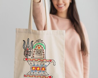 May include: Beige tote bag featuring a colorful illustration of a religious figure with the text "Shiva is Auspicious" and "Shiva Shiva Shiva" in red. The design includes a trident and other symbolic elements.