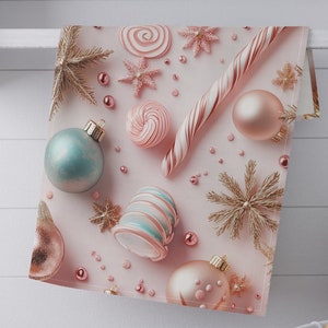 May include: A pink and white Christmas-themed kitchen towel featuring ornaments, candy canes, and snowflakes. The towel has a light pink background with gold, pink, and blue decorative elements. The towel is hanging on a white shelf.