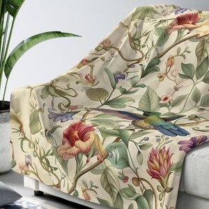 Peut inclure: Une couverture en jet de couleur crème avec un motif floral représentant un colibri, des fleurs et des feuilles. La couverture est jetée sur un canapé blanc.