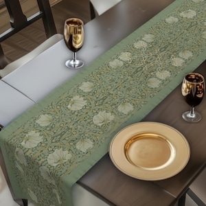 May include: A dining table set for a meal, featuring a sage green floral table runner. The table is set with gold-rimmed plates, wine glasses, and a bowl of decorative eggs. A fireplace and window with flowers are in the background.