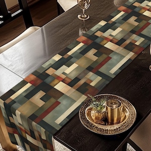 May include: A rectangular table runner with a geometric pattern in shades of brown, beige, and blue. The runner is placed on a dark wooden table, with chairs and glassware visible. The table is set for a meal.
