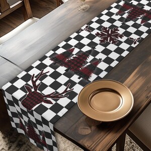May include: A table runner with a black and white chequered pattern. The runner has red and black plaid deer, trees, and snowflake designs. It's on a wooden table with a gold plate and glasses. The table is surrounded by wooden chairs.