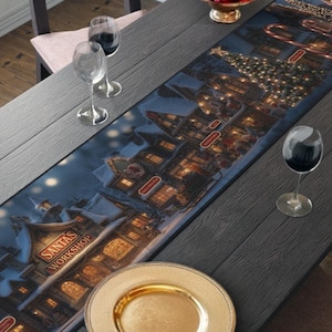 May include: A Christmas-themed table runner featuring a detailed illustration of Santa's Workshop. The runner is predominantly blue with a festive village scene, including buildings, lights, and the text "Santa's Workshop". A gold plate and wine glasses are on the table.