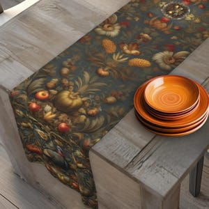 May include: A dark floral table runner with a pattern of flowers, pumpkins, and fruit. The runner is paired with a stack of orange ceramic plates. The table setting is on a rustic wooden table.