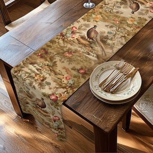 May include: A long rectangular table runner featuring a repeating pattern of pheasants, green foliage, and pink and yellow roses on a beige background. A stack of two white ceramic plates with gold rims and three gold-toned forks are placed on top.