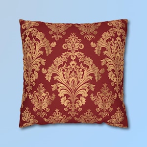 May include: A square throw cushion featuring a deep red background with a repeating gold damask pattern. The intricate, symmetrical floral and leaf motifs are printed in a metallic gold tone, covering the entire front surface of the cushion.