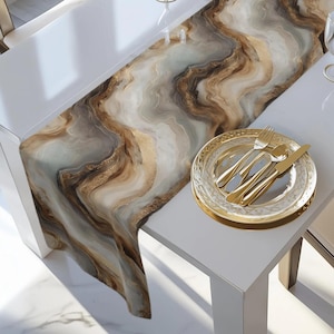 May include: A table setting featuring a decorative table runner with a marble design in shades of brown, gold, and white. The table is set with gold-colored cutlery, plates, and a glass dish.