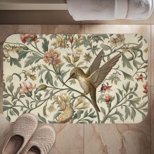 May include: Rectangular bath mat featuring a hummingbird with outstretched wings, perched amidst a floral design. The mat's background is cream-colored, with a pattern of flowers and leaves in shades of green, pink, and yellow. A pair of beige slippers are in the foreground.
