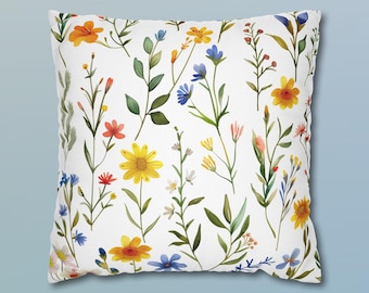 May include: A square throw pillow featuring a white background with a repeating pattern of various wildflowers. The botanical print includes yellow daisies, blue cornflowers, orange blossoms, and green stems with leaves scattered across the surface.