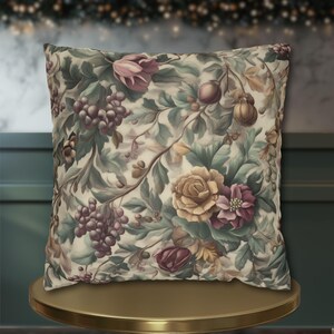 May include: Square decorative pillow with a floral pattern featuring grapes, roses, and foliage in shades of purple, yellow, and green. The pillow has a cream-colored background and is displayed on a gold-colored surface.