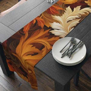 Peut inclure: Chemin de table décoratif avec un motif répété de feuilles stylisées dans les tons de marron, orange et crème. Le chemin de table est posé sur une table en bois foncé, avec des assiettes blanches et des couverts.