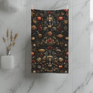 May include: A decorative towel with a dark background featuring a repeating pattern of red apples, gold-colored ornaments, and nutcracker figures. The towel is hanging on a clothesline against a white tiled wall.